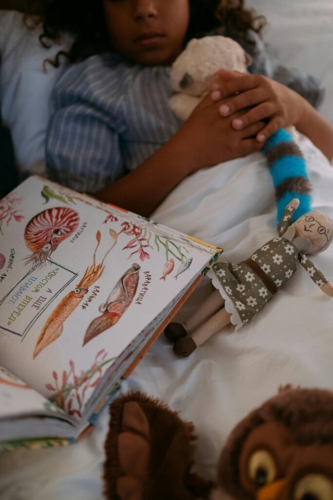 A child lies in bed holding a stuffed toy, surrounded by a book and other toys.